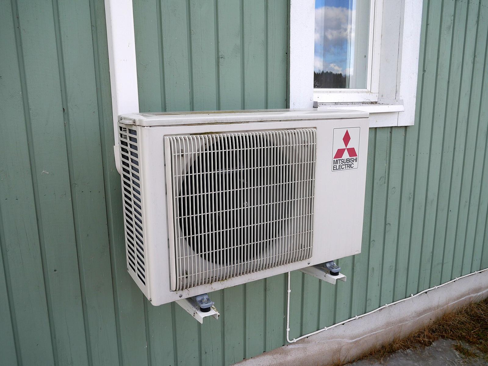 Witte buitenunit van een airco op een muurbeugel onder een raam, tegen een groengeverfde houten gevel