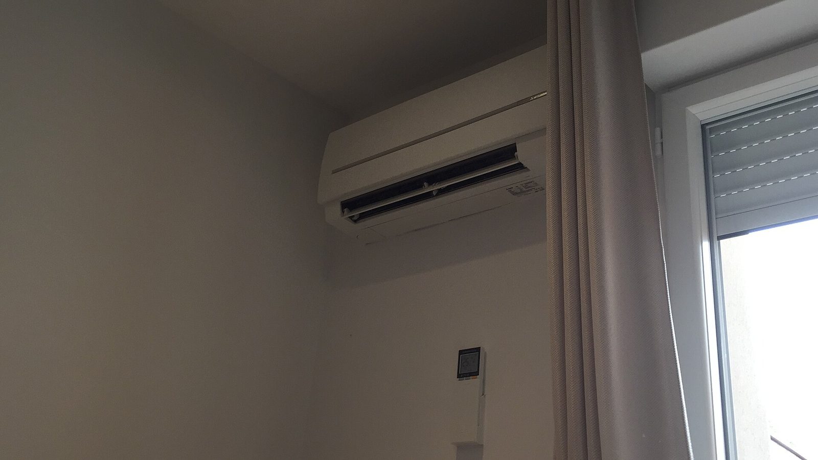 Witte binnenunit van een split-airco hoog aan de muur naast een raam met grijs gordijn, met een bedieningspaneel eronder