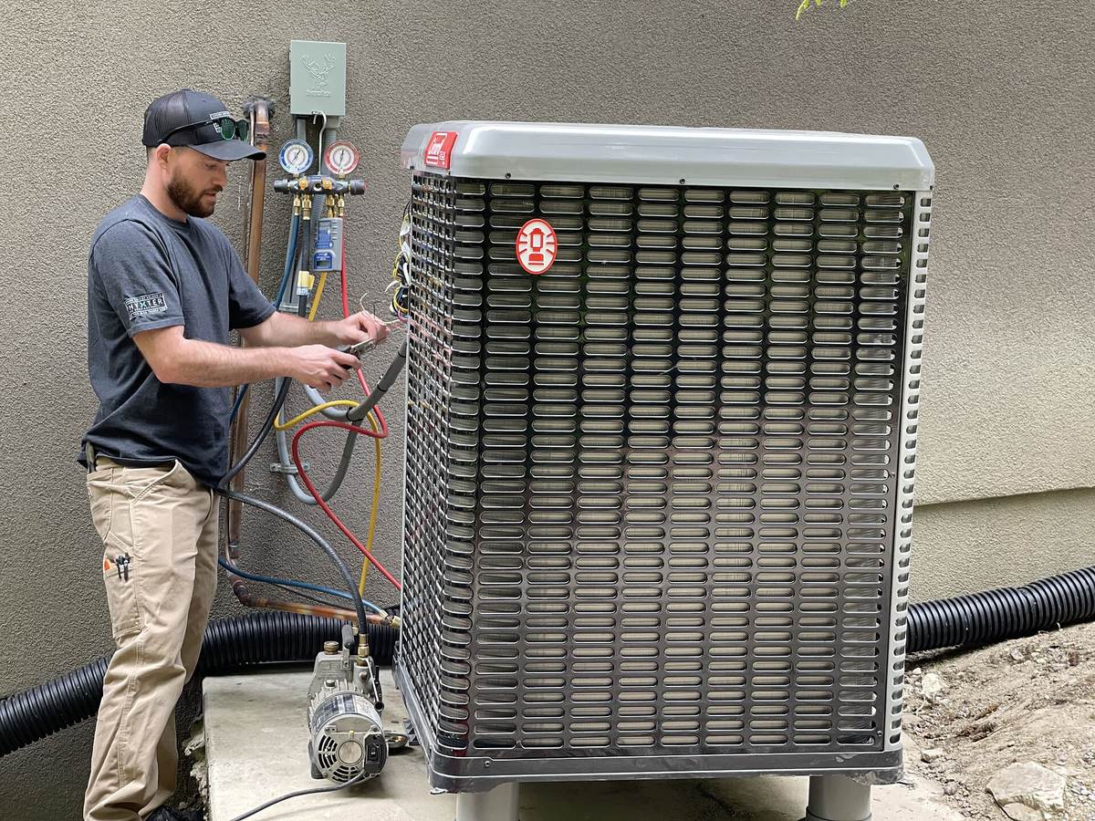 Technicus sluit slangen met manometers aan op de buitenunit van een warmtepomp naast een grijze gevel, met een vacuümpomp