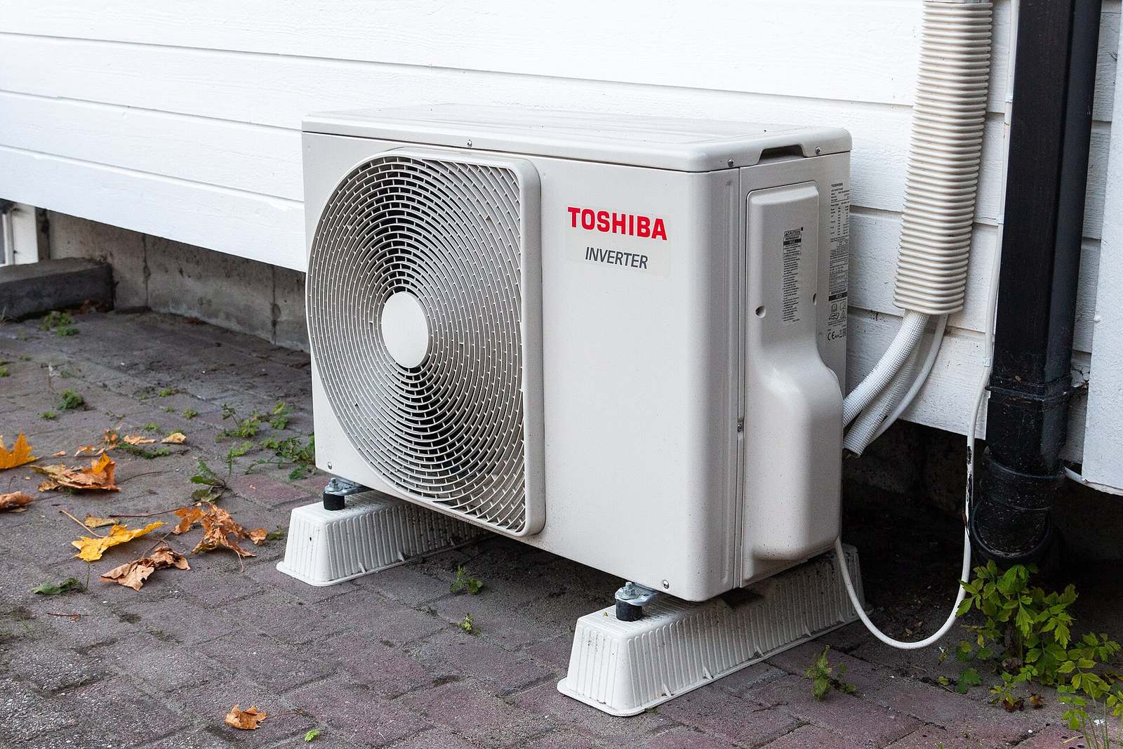 Witte buitenunit van een airco op kunststof voetjes op een klinkervloer tegen een witte houten gevel, met herfstbladeren